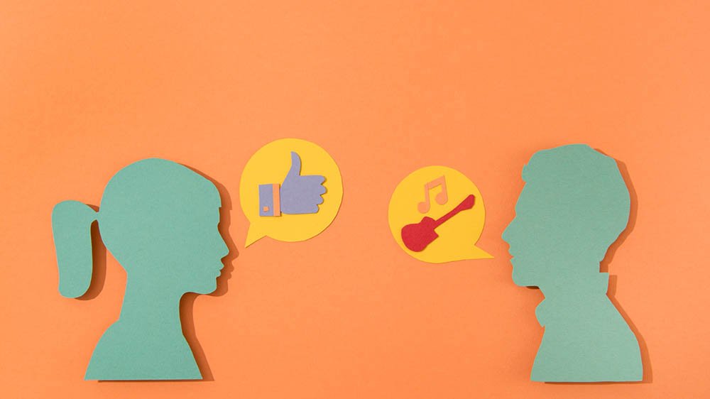 “Two paper-cut silhouettes facing each other against an orange background. The left silhouette has a ponytail and a speech bubble with a thumbs-up icon. The right silhouette has short hair and a speech bubble containing a guitar and musical note icon.”