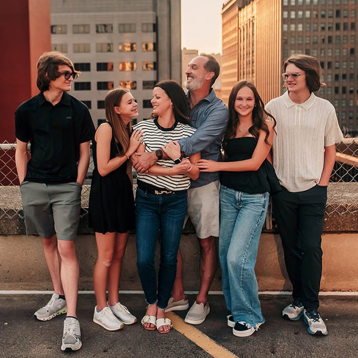 Six casually dressed people—three males and three females—walk together on a rooftop parking lot in St. Louis, Missouri. They appear relaxed and cheerful, enjoying each other’s company. The Gateway Arch and tall buildings rise in the background, with warm lighting suggesting early morning or late afternoon.