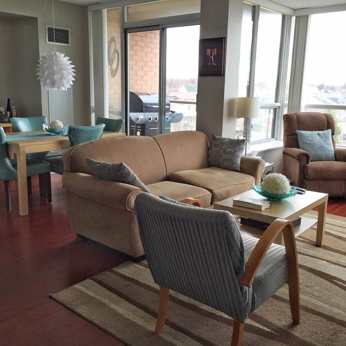 A spacious, sunlit living and dining area with large windows and sliding glass doors. The living room includes a beige sectional sofa with patterned pillows, a striped armchair, and a recliner with a light blue pillow. A light wood coffee table holds a decorative bowl of white flowers and books. The dining area features a wooden table with teal upholstered chairs and a modern white pendant light overhead. Outside, a barbecue grill is visible on the balcony. Hardwood floors and a striped area rug enhance the cozy, welcoming atmosphere.