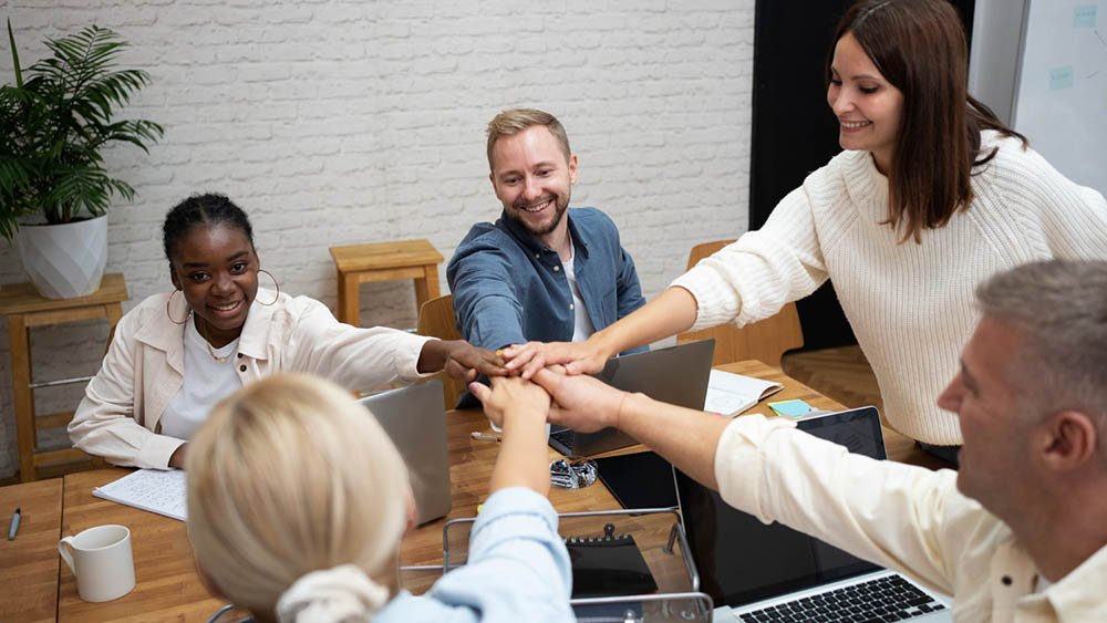 “Group of five people gathered around a wooden table in a modern office, placing their hands together in the center as a team-building gesture. They are smiling and surrounded by laptops, notebooks, pens, and coffee mugs. A potted plant and stools are visible in the background, with bright lighting contributing to a collaborative atmosphere.”2