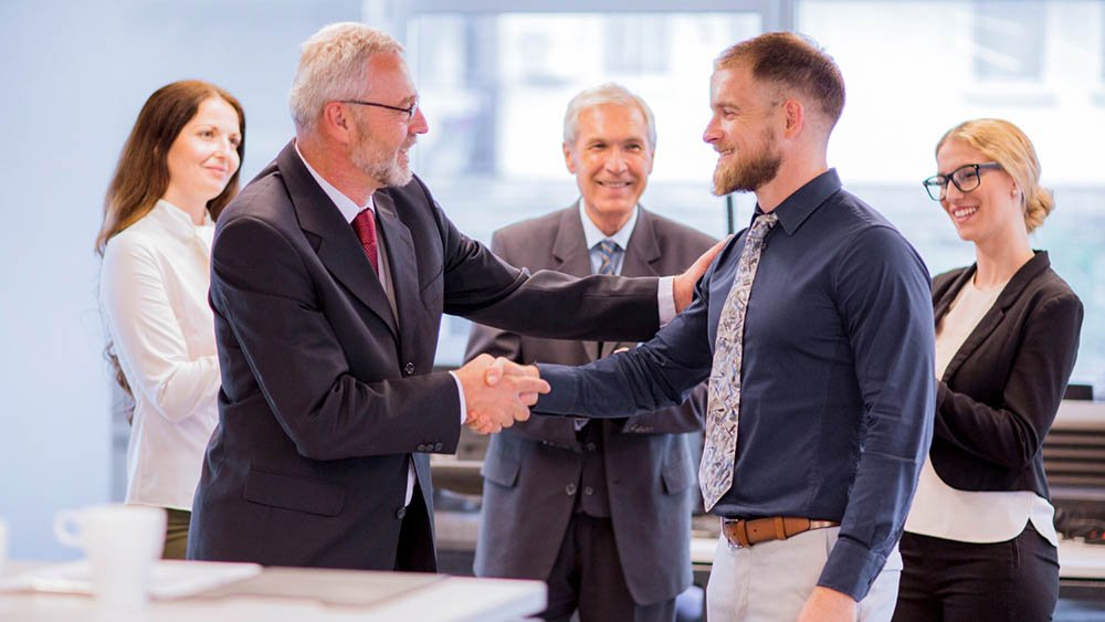 “Five individuals in business attire gathered in a professional office setting with large windows and office furniture. Two men are shaking hands, with one placing a hand on the other’s shoulder in a congratulatory gesture. The other three people stand nearby, observing with positive expressions.”