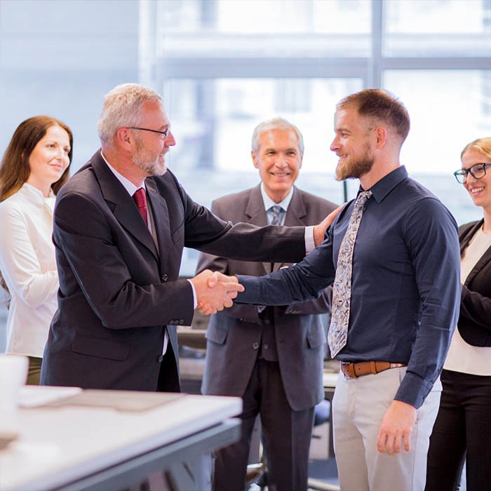 “Five individuals in business attire gathered in a professional office setting with large windows and office furniture. Two men are shaking hands, with one placing a hand on the other’s shoulder in a congratulatory gesture. The other three people stand nearby, observing with positive expressions.”