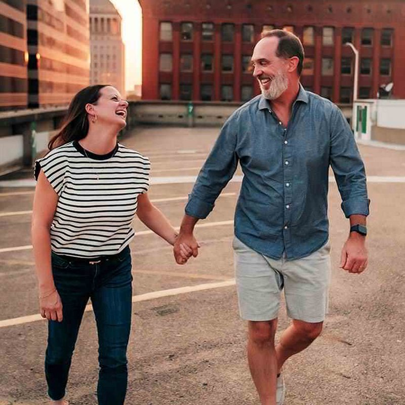 Two people walk hand-in-hand across the top level of an urban parking garage, surrounded by tall buildings including a red brick structure and modern glass towers. The warm light of early morning or late afternoon casts a soft glow over the scene. They face each other, sharing a quiet, intimate moment amid the cityscape.
