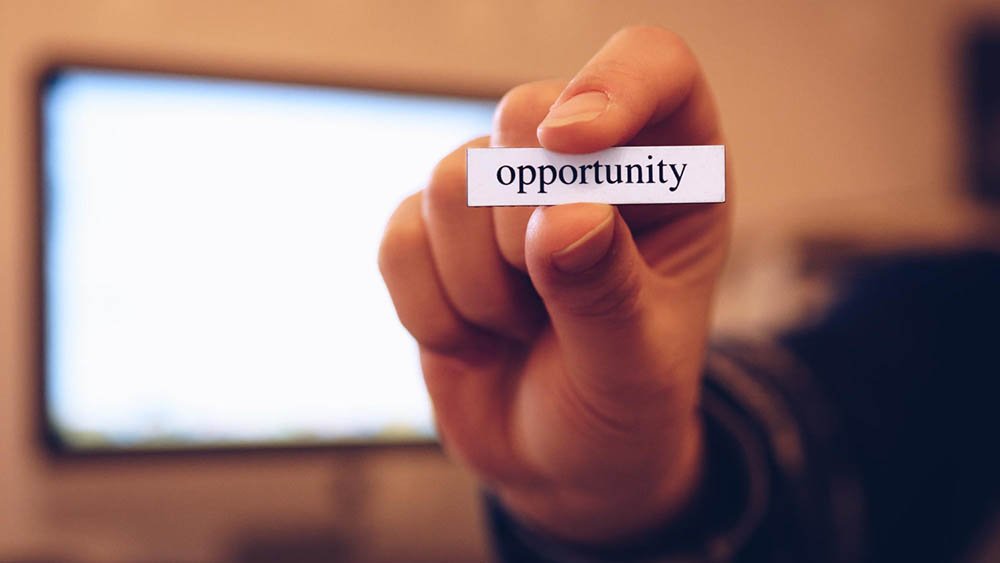 A close-up of a hand holding a small rectangular paper with the word “opportunity” printed in bold black letters. The background is softly blurred, showing a computer monitor in an indoor workspace. The image conveys a symbolic message of motivation and potential.