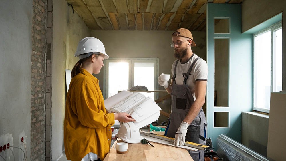 “Two individuals in a construction setting reviewing a large architectural blueprint. One wears a white hard hat and yellow shirt, the other wears overalls and a cap while holding a cup. They stand in a room with exposed brick, unfinished walls, and construction tools on a table, lit by natural light from nearby windows.”
