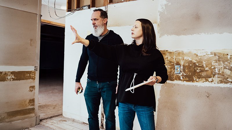 Two people stand in a partially renovated room with unfinished walls. One person holds a tablet and gestures toward the space, while the other holds a tape measure and listens. Exposed drywall, patches on the wall, and an electrical outlet indicate ongoing construction or planning.
