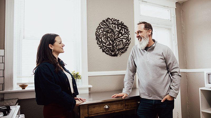 Two people stand in a kitchen having a conversation. One leans against a wooden counter with drawers, while the other faces them. The room has neutral walls with white trim, two windows with blinds, and a decorative metal wall art piece between them. A small plant sits on the counter.
