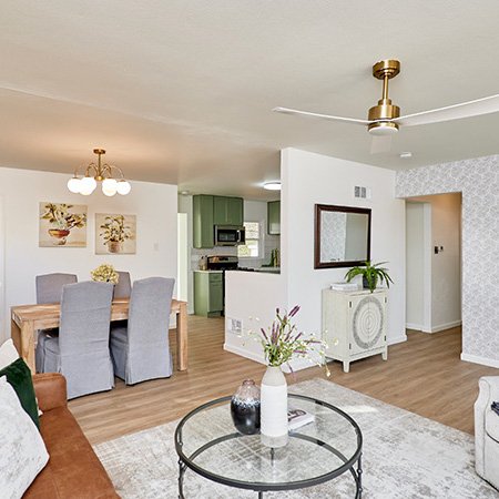 A modern living and dining area features a brown leather sofa with green and white pillows, a round glass coffee table with decorative vases and flowers, and a gray armchair with a dark throw blanket. A ceiling fan hangs above the seating area. Behind it, a wooden dining table with six light gray upholstered chairs sits beneath a chandelier. The kitchen is visible in the background with green cabinets and stainless steel appliances. The room includes white walls, a geometric accent wall, framed artwork, and a white cabinet with a mirror and plant.