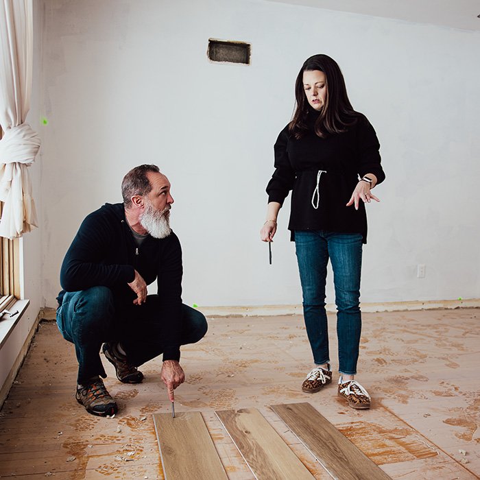 A man crouches on the floor of a room under renovation, pointing at several wooden floor planks laid out in front of him. A woman stands beside him holding a tool and gesturing toward the planks. The room has bare walls and an unfinished floor, indicating that flooring installation is in progress