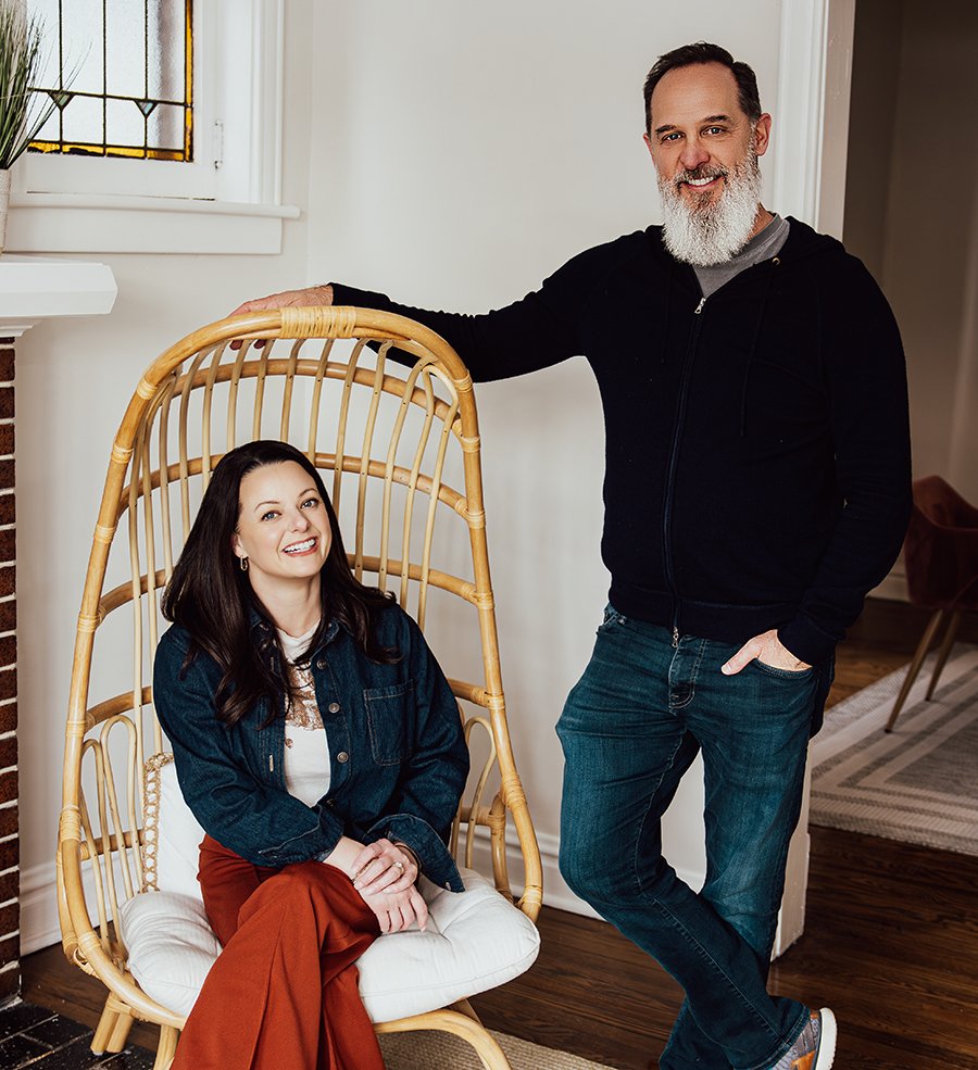 MATT AND REBEKA HERO SHOT Two adults are in a cozy living room. One person sits in a wicker egg chair with a white cushion, wearing a denim jacket and rust-colored pants. The other person stands beside the chair, resting a hand on it, dressed in a black zip-up hoodie and jeans. Behind them is a white wall with a stained-glass window, a brick fireplace, and an open doorway leading to another room with a rug and chair.