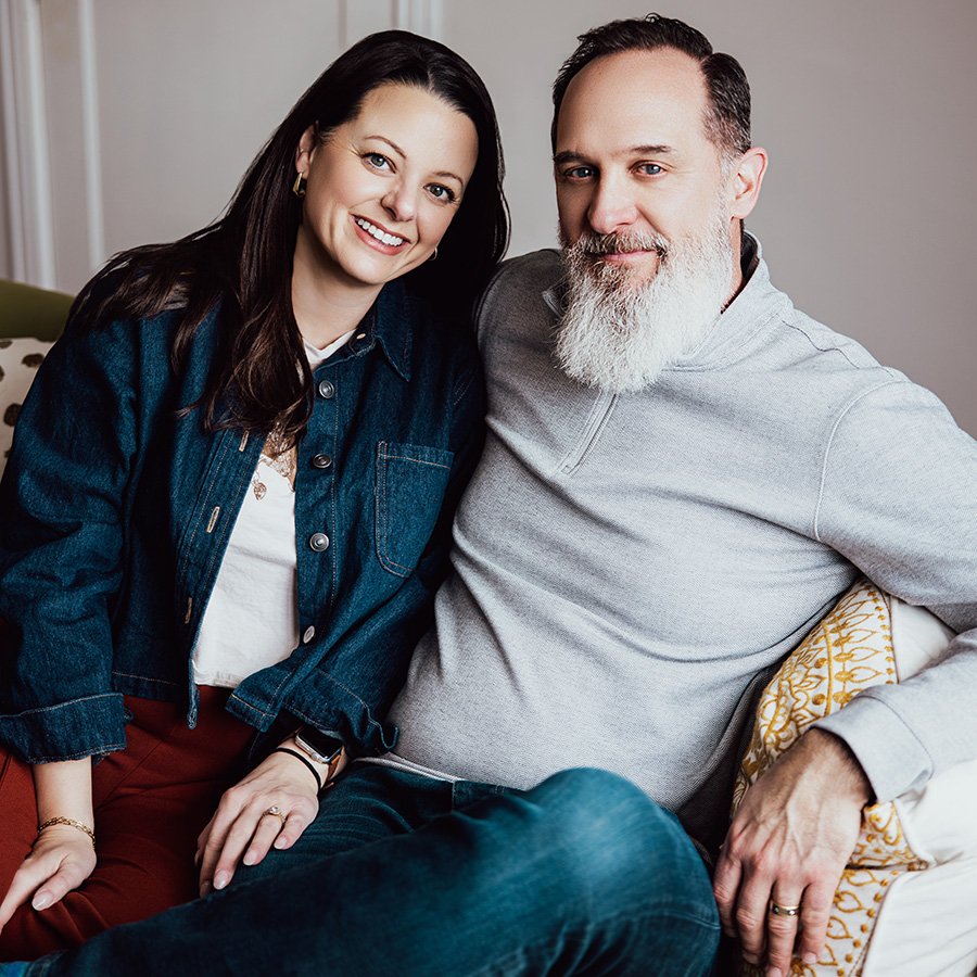 MATT REB PROFILE “Two people sit close together on a white couch. The person on the left has long dark hair and wears a denim jacket over a white top with red pants. The person on the right has a full white beard and wears a light gray pullover with blue jeans. A yellow patterned pillow rests beside them, and the indoor setting is softly lit with a neutral background.”