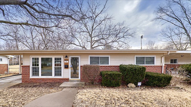 A single‑story mid‑century modern house with red brick and beige siding is shown from the front. Three large windows sit on the left side, with two smaller horizontal windows on the right. A glass front door with the house number 303 is centered above a small concrete step. The yard has dry grass, trimmed bushes, and leafless shrubs, with bare trees and a partly cloudy sky in the background.