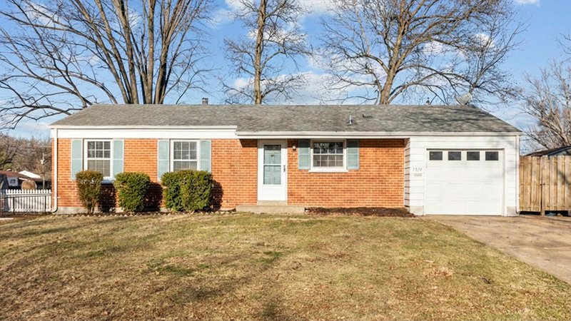 A single‑story brick house with a gray shingled roof and white trim is shown from the front. Light blue shutters frame the windows, and a central door sits above a small concrete stoop. Four neatly trimmed bushes line the front of the house, with a dry lawn and leafless trees in the background. The house number 2291 appears beside the garage door.